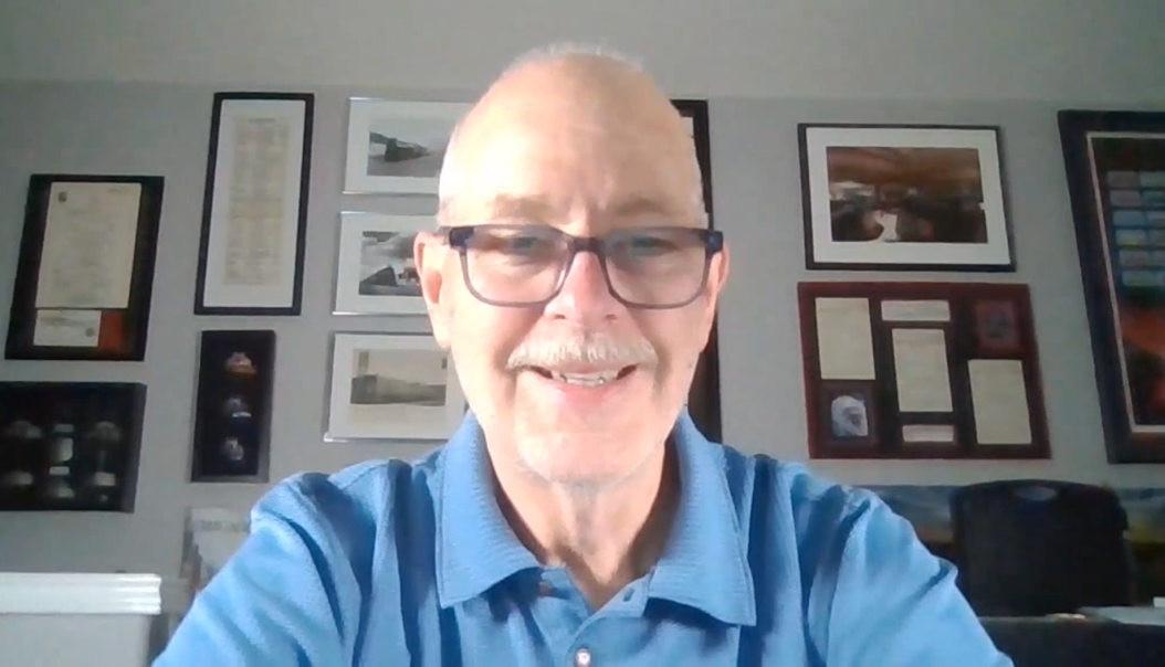 Smiling man with glasses and a blue polo shirt on a video call, seated before a wall of framed photos and documents.